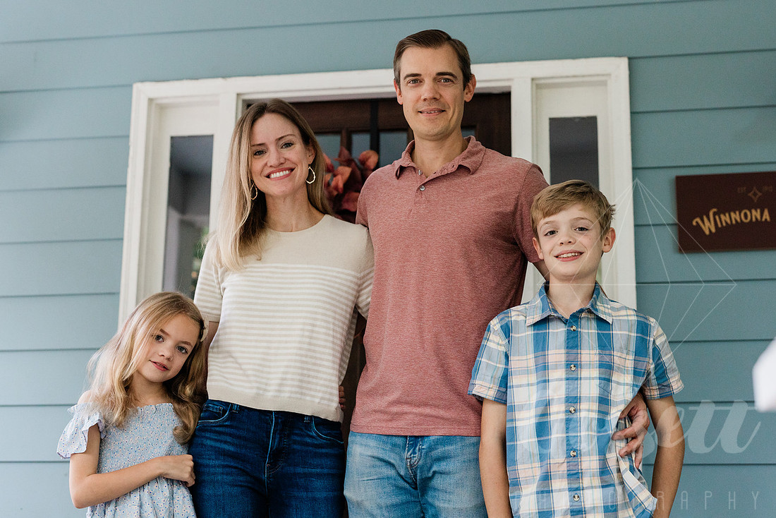 Family of four sitting on their front porch steps, smiling and holding hands in golden afternoon light.