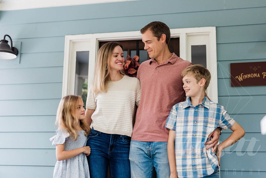 Family of four sitting on their front porch steps, smiling and holding hands in golden afternoon light.