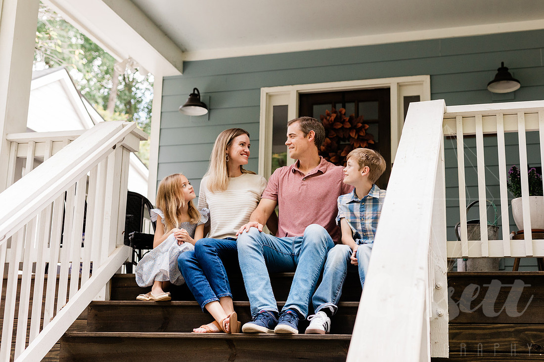 Family of four sitting on their front porch steps, smiling and holding hands in golden afternoon light.