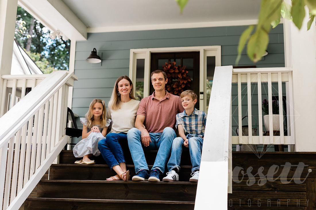 Family of four sitting on their front porch steps, smiling and holding hands in golden afternoon light.