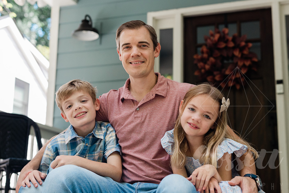 Family of four sitting on their front porch steps, smiling and holding hands in golden afternoon light.