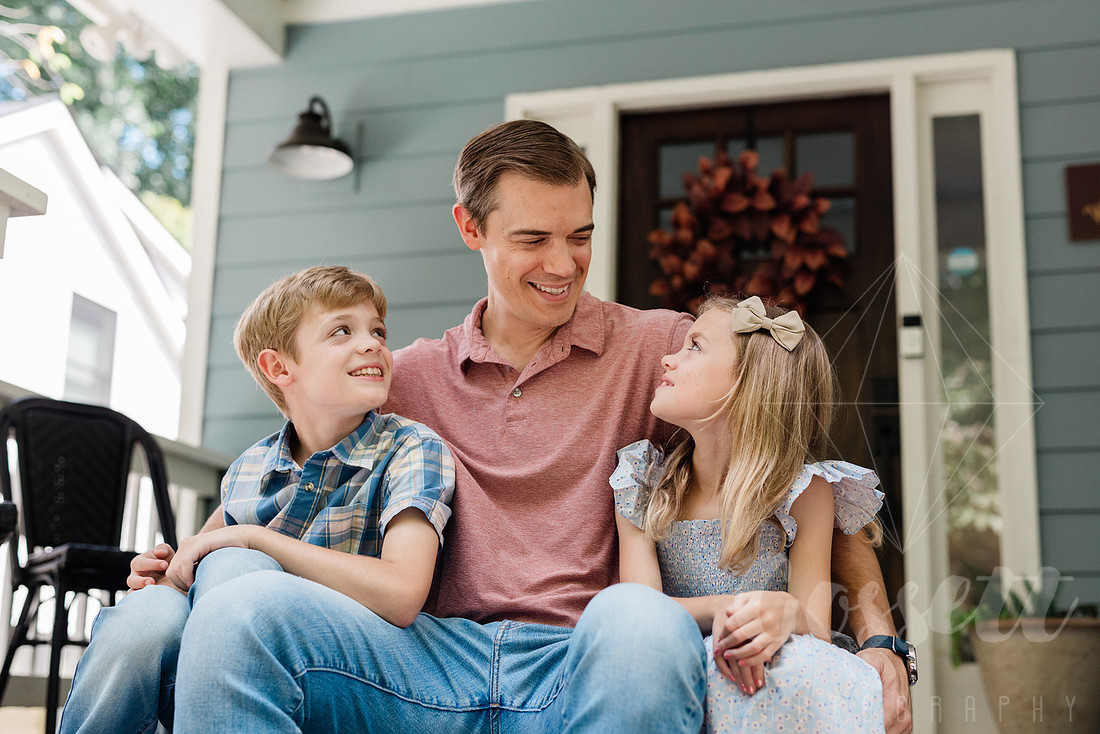 Family of four sitting on their front porch steps, smiling and holding hands in golden afternoon light.