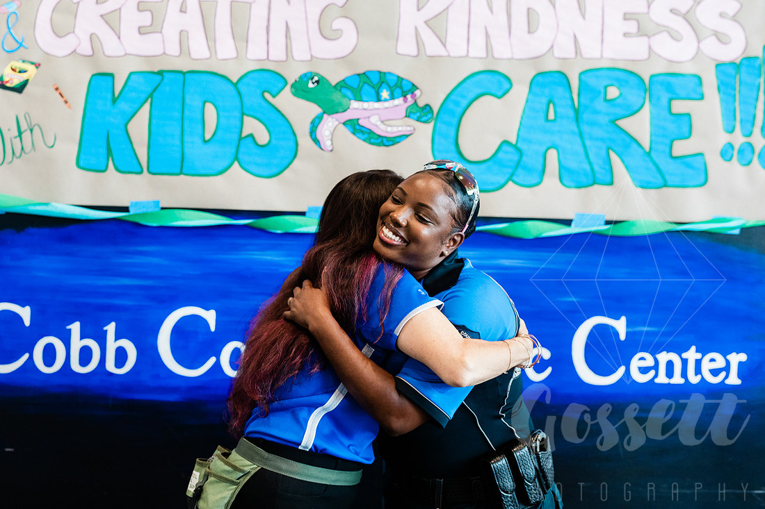 Police Officer and CEO of KIDS CARE, Aimee Burke, hug in front of colorful sign at the School Supply Drive.